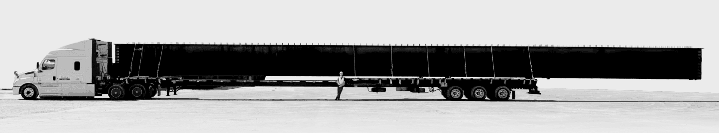 Panoramic black and white photo of a semi-truck hauling a long industrial load, with a crew member for scale